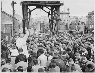 Photo Credit: US National Archives. Father (Major) Edward J. Waters, Catholic Chaplain from Oswego, New York, conducts Divine Services on a pier for members of the first assault troops thrown against Hitler's forces on the continent. Weymouth, England. 06/06/1944.